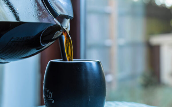 hot dark filter coffee being poured from a glass carafe into a black coffee cup or mug. patio doors leading to a garden in the background.